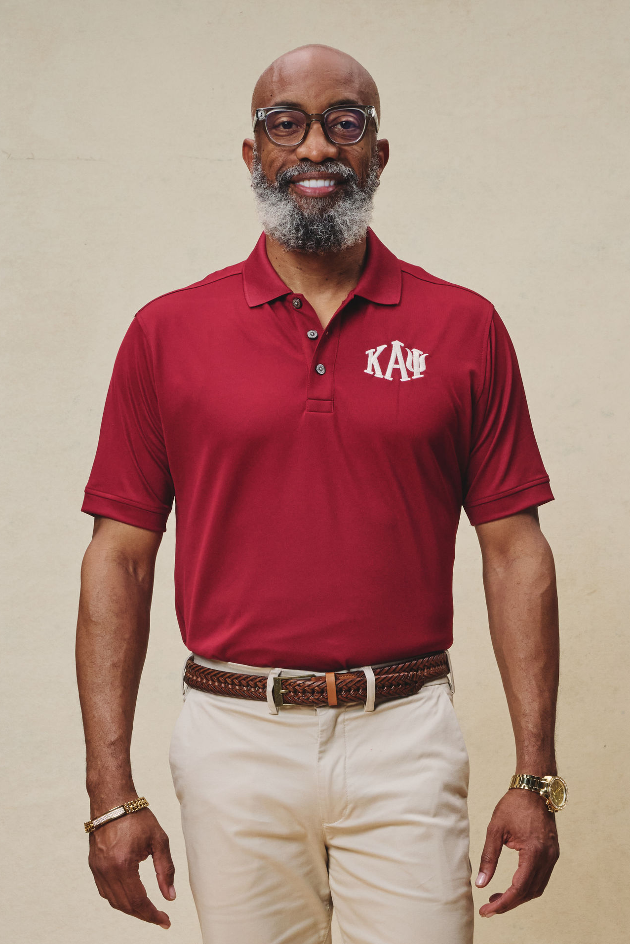 A man wearing a red short-sleeved polo shirt with the Kappa Alpha Psi fraternity logo on the chest, paired with beige pants and a brown belt.