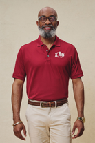 A man wearing a red short-sleeved polo shirt with the Kappa Alpha Psi fraternity logo on the chest, paired with beige pants and a brown belt.
