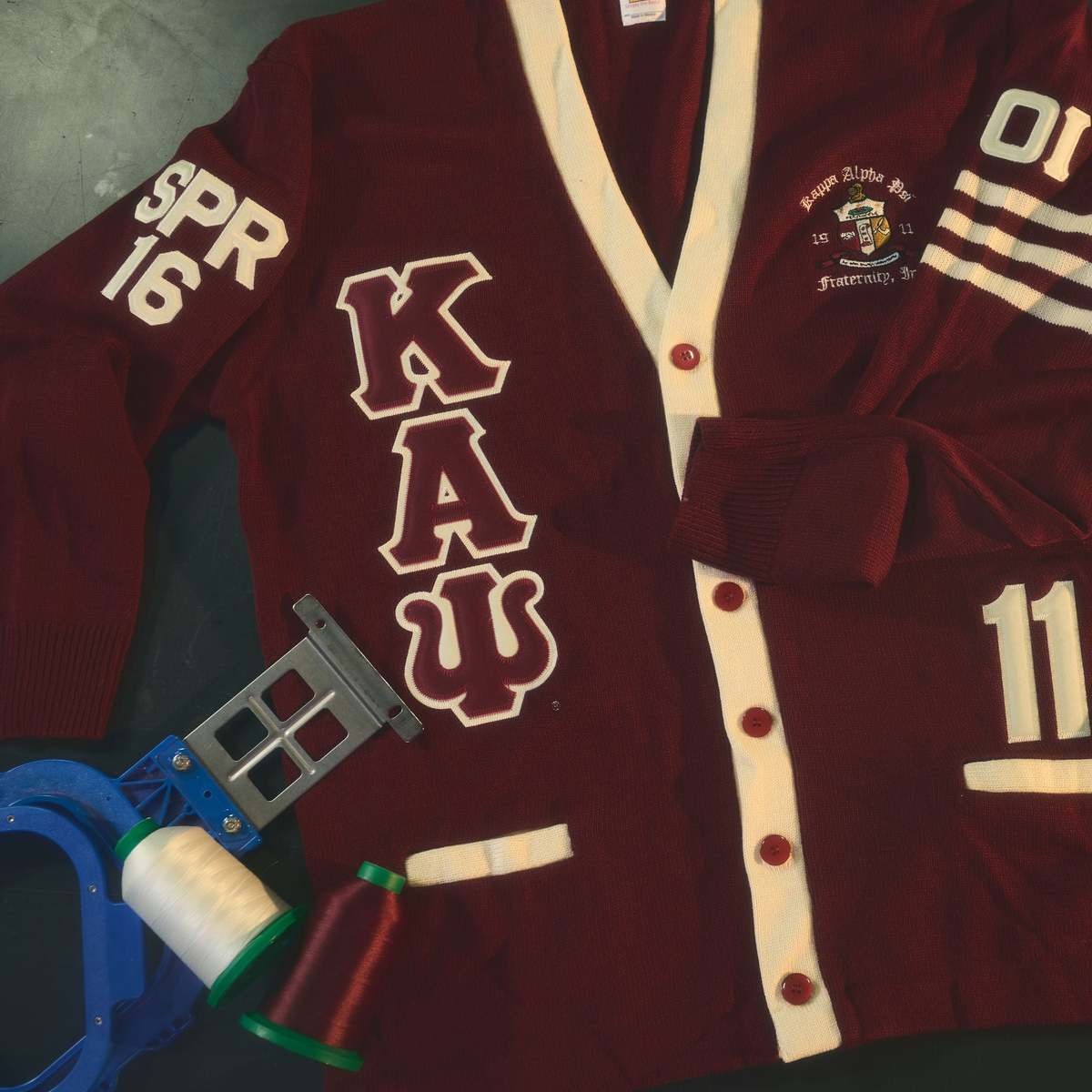 A maroon varsity-style cardigan with cream trim and embroidered details, laid flat on a dark surface with matching decorative patches and small props around it. The cardigan shows large Greek letters, numbers, and a crest patch on the chest.